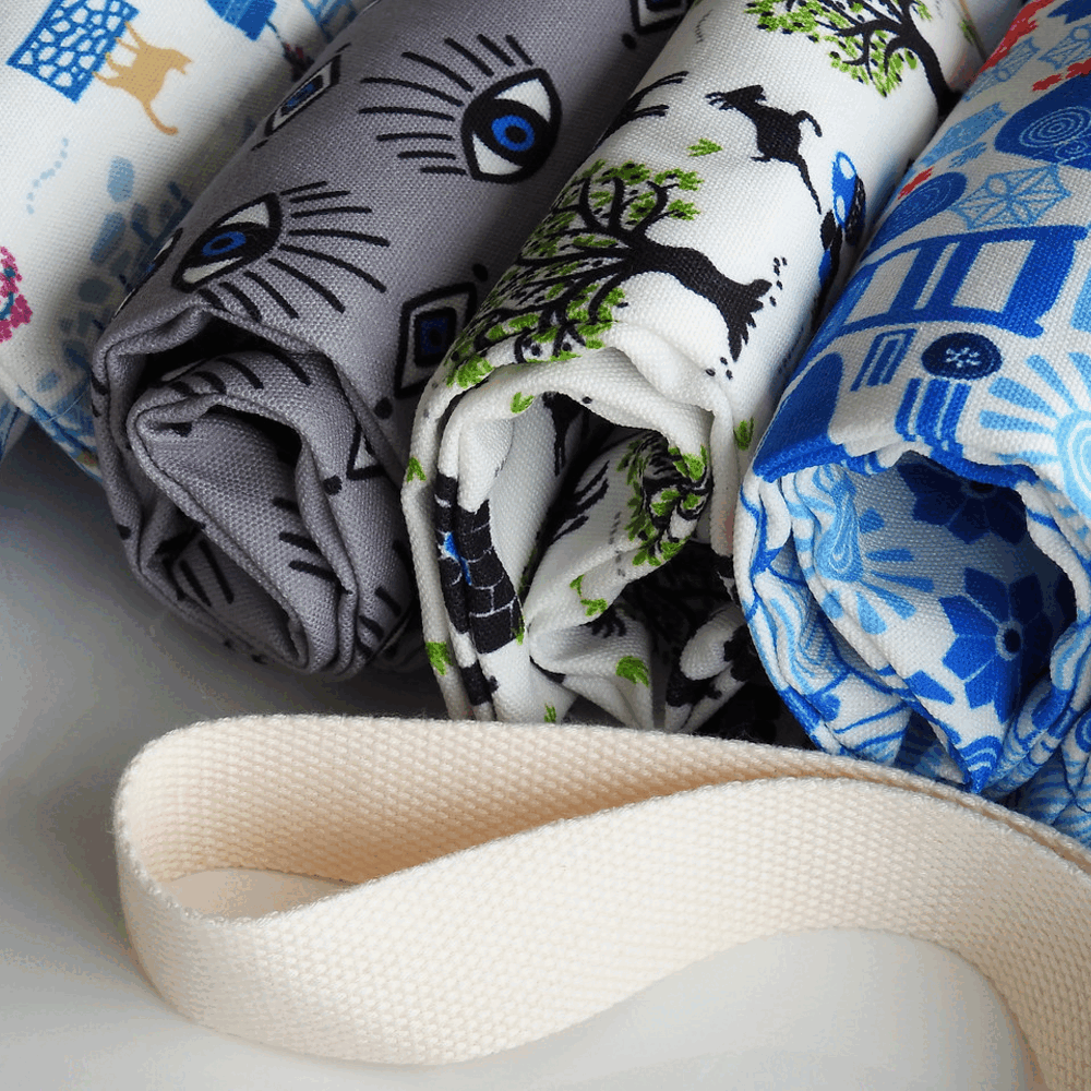 Three rolled fabric items with printed patterns in gray, white, and blue, arranged together with a cream-colored woven strap in the foreground.
