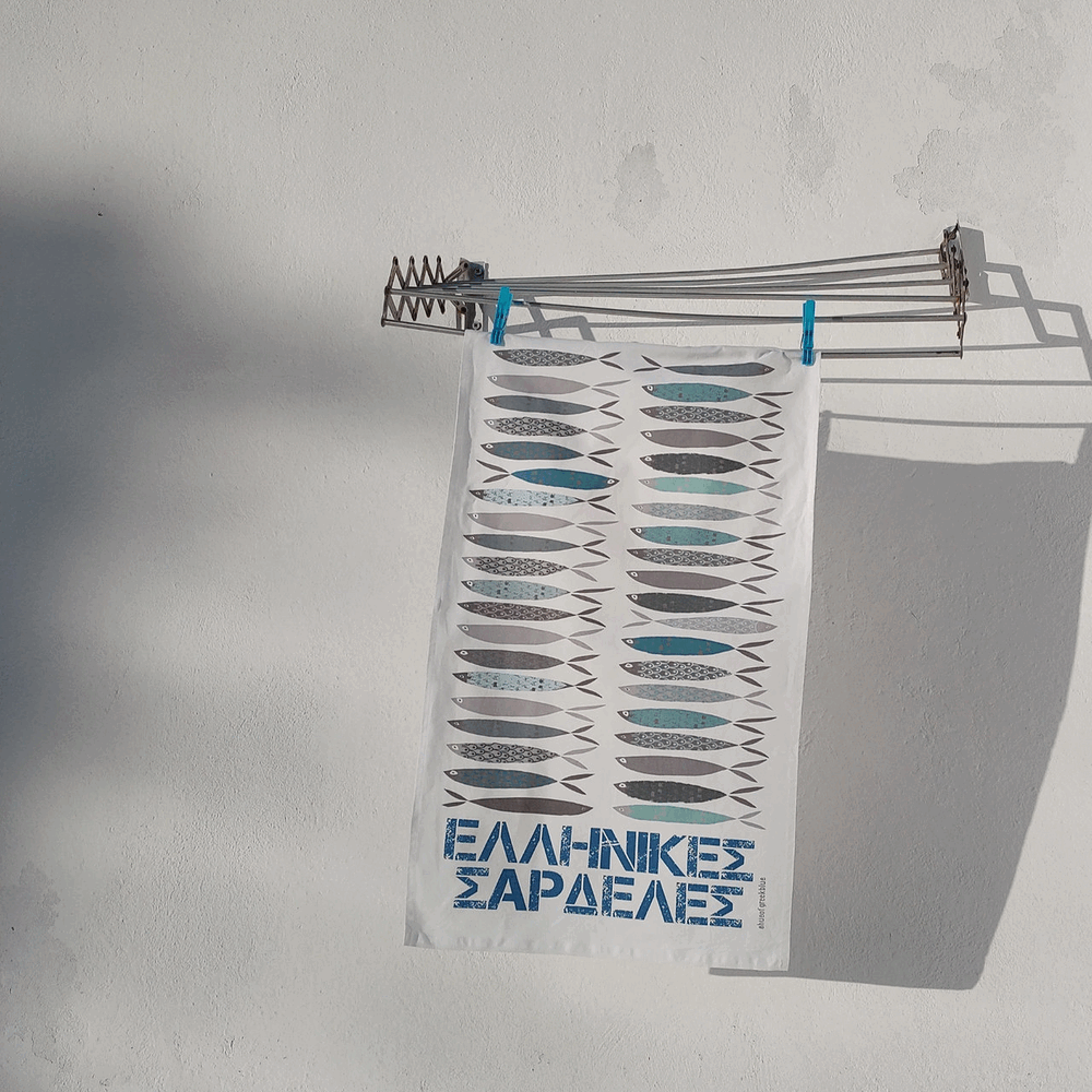 A white hanging textile banner or flag with rows of gray and blue fish illustrations and the text 'ΕΑΒΕΛΩΑΒΕ ΓΑΒΕΑΚΑΞ' at the bottom, clipped to a metal clothesline against a white wall.