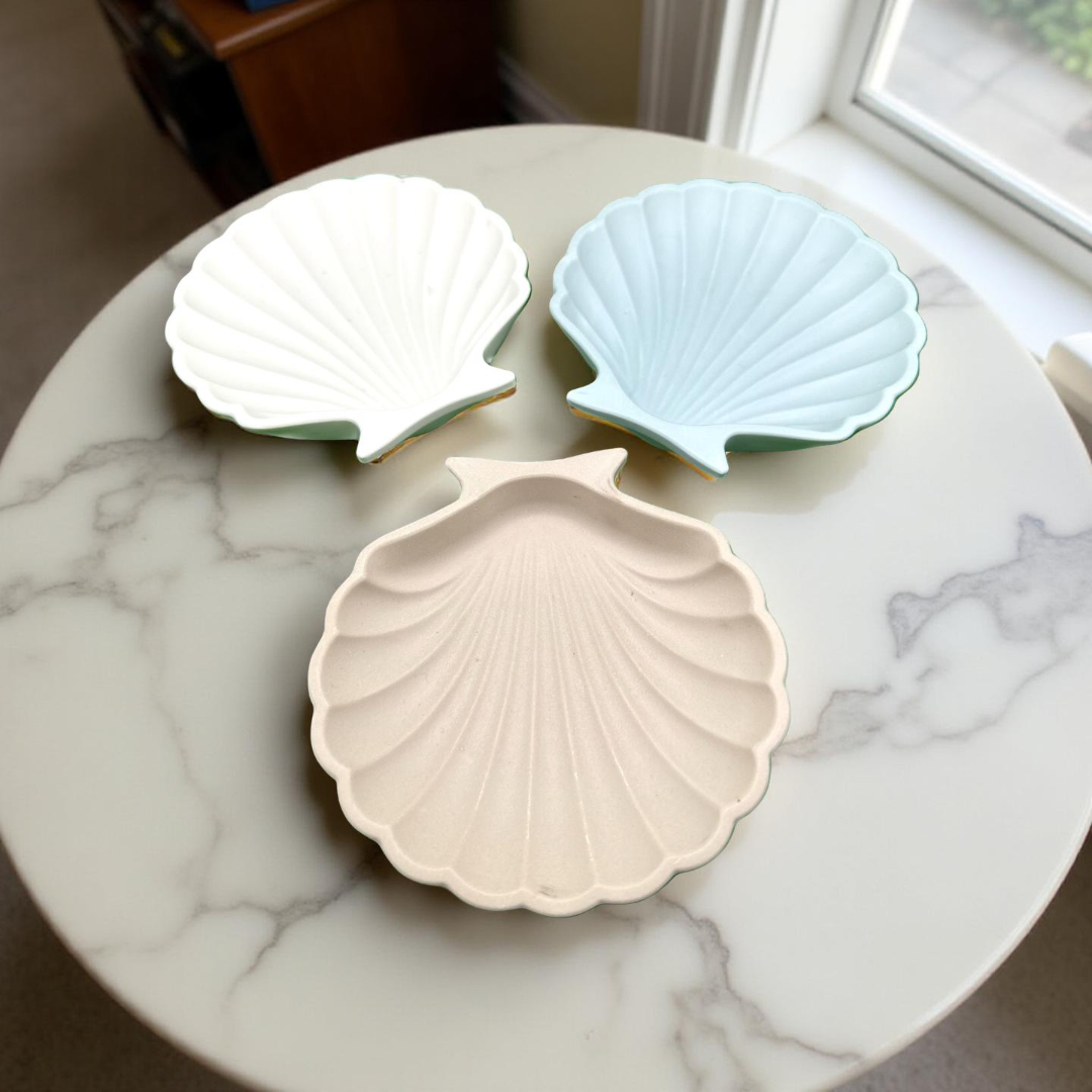 Three shell-shaped ceramic dishes in white, light blue, and beige on a marble surface.
