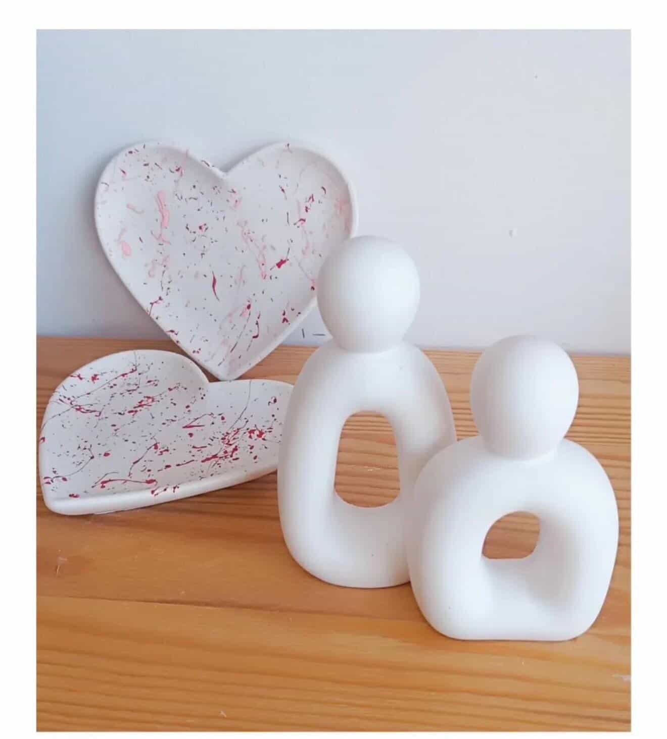 Two white abstract sculptures on a wooden surface with two heart-shaped objects in the background.