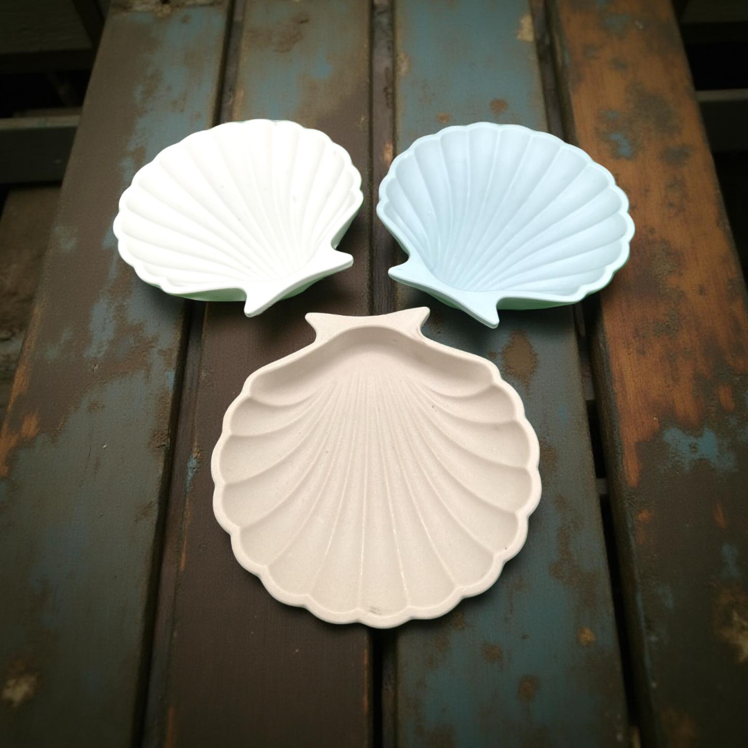 Three shell-shaped dishes in white, light blue, and beige on a rustic wooden surface.