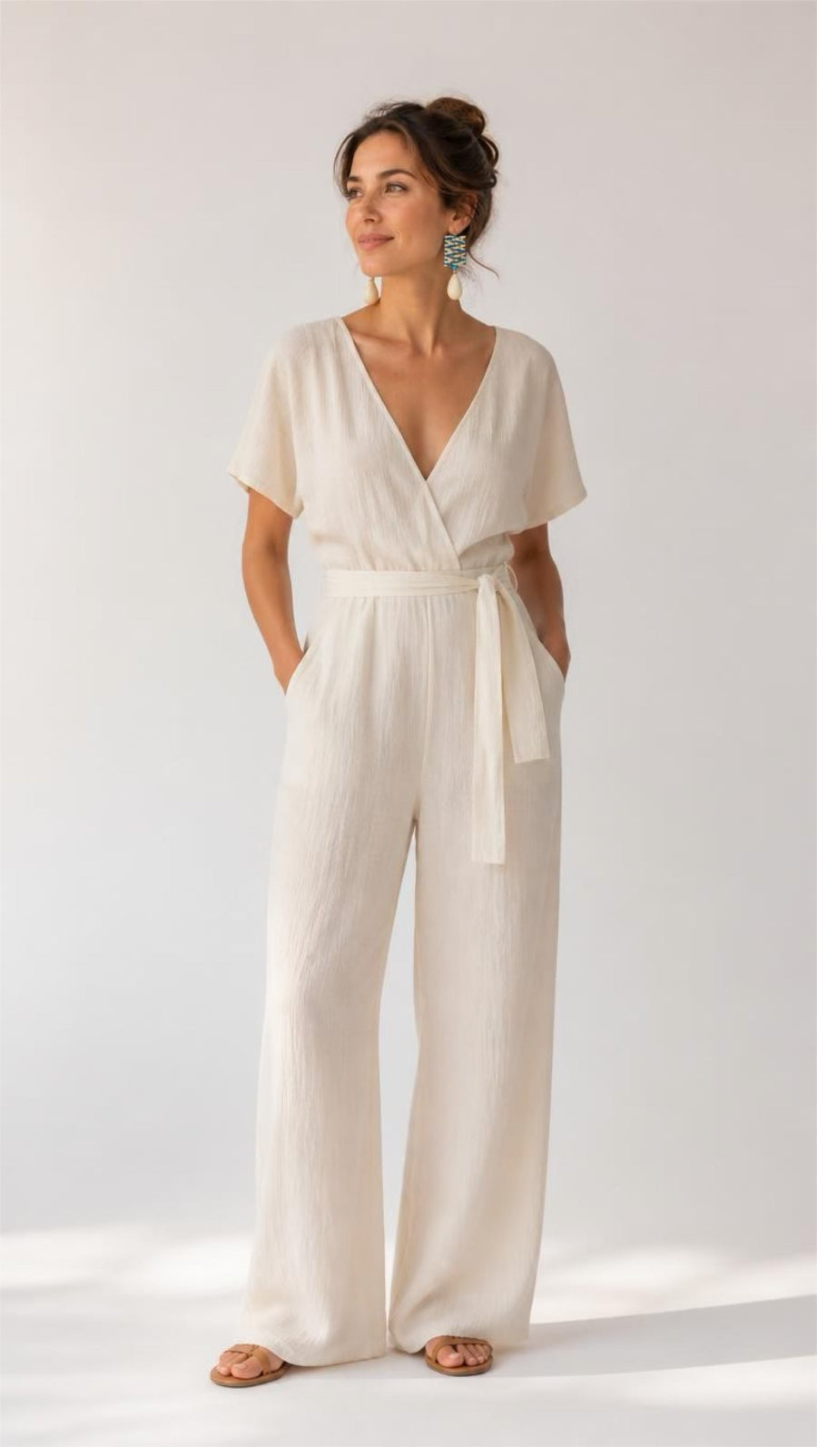 A woman modeling a cream-colored linen jumpsuit with short sleeves, a wrap-style V-neck, a matching fabric tie belt at the waist, and wide legs, standing against a plain white studio background.