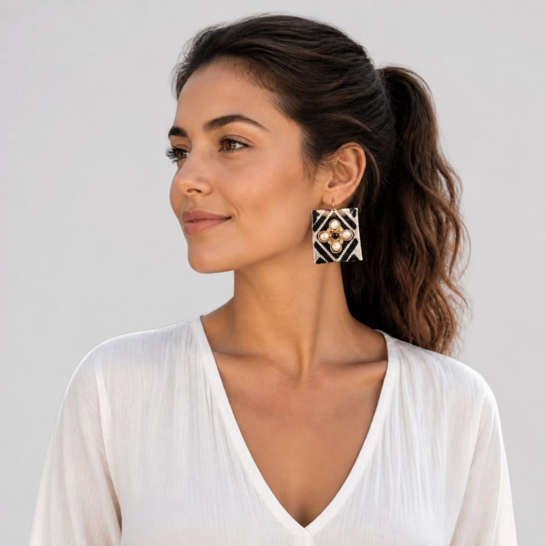 A woman with her hair in a ponytail wearing a white V-neck top and a geometric black, white, and gold statement earring, shown against a light gray studio background.