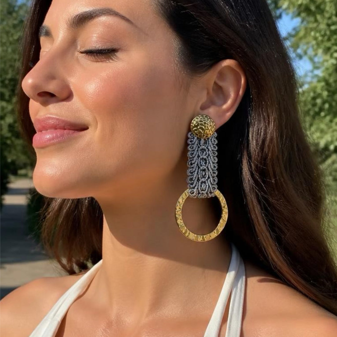 A woman outdoors wearing a large statement earring with a textured gold top, a long silver and gold woven drop, and a large gold ring.