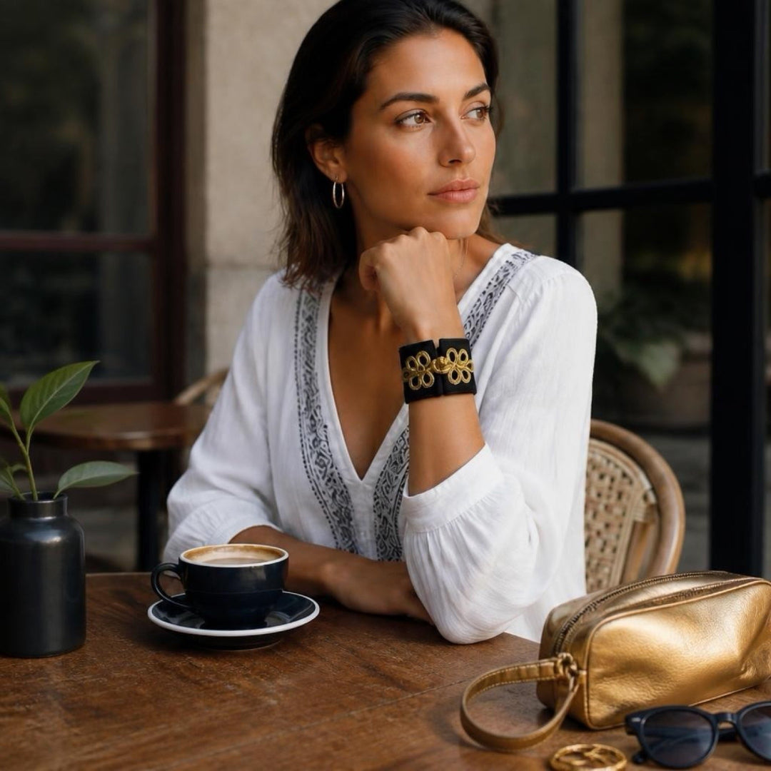 A woman in a white blouse sits at a wooden cafe table wearing a black cuff bracelet with gold geometric patterns. A coffee cup, a small black vase with a plant, and a metallic gold handbag are on the table next to her.