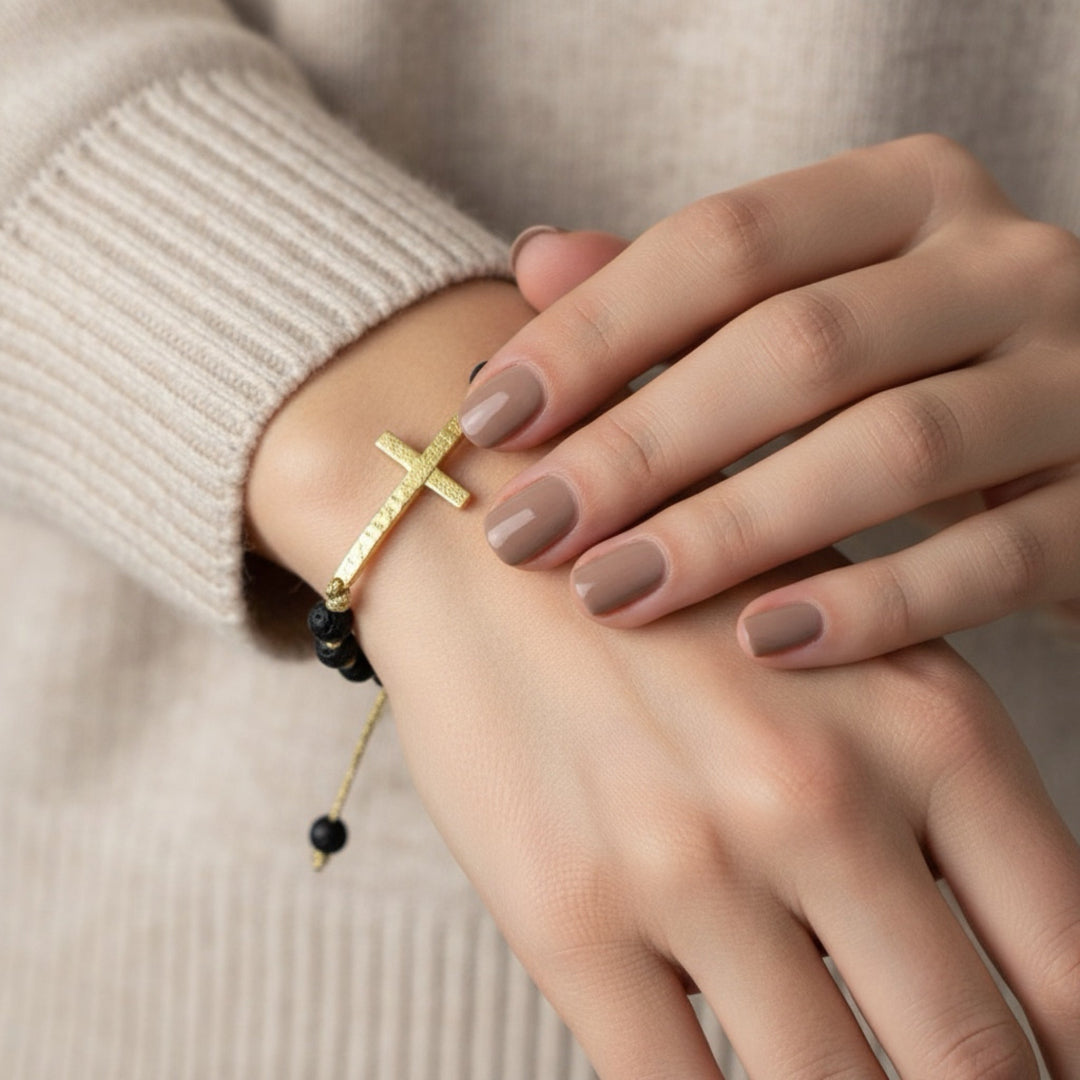Close-up of a hand wearing a gold bracelet with a cross charm on a neutral background