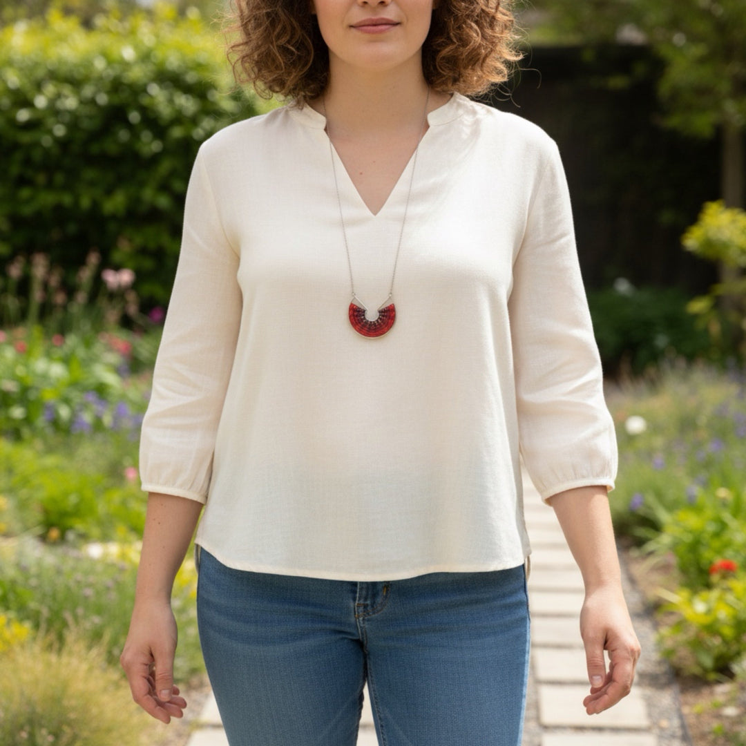 Woman wearing a white blouse and blue jeans with a red necklace, standing in a garden.