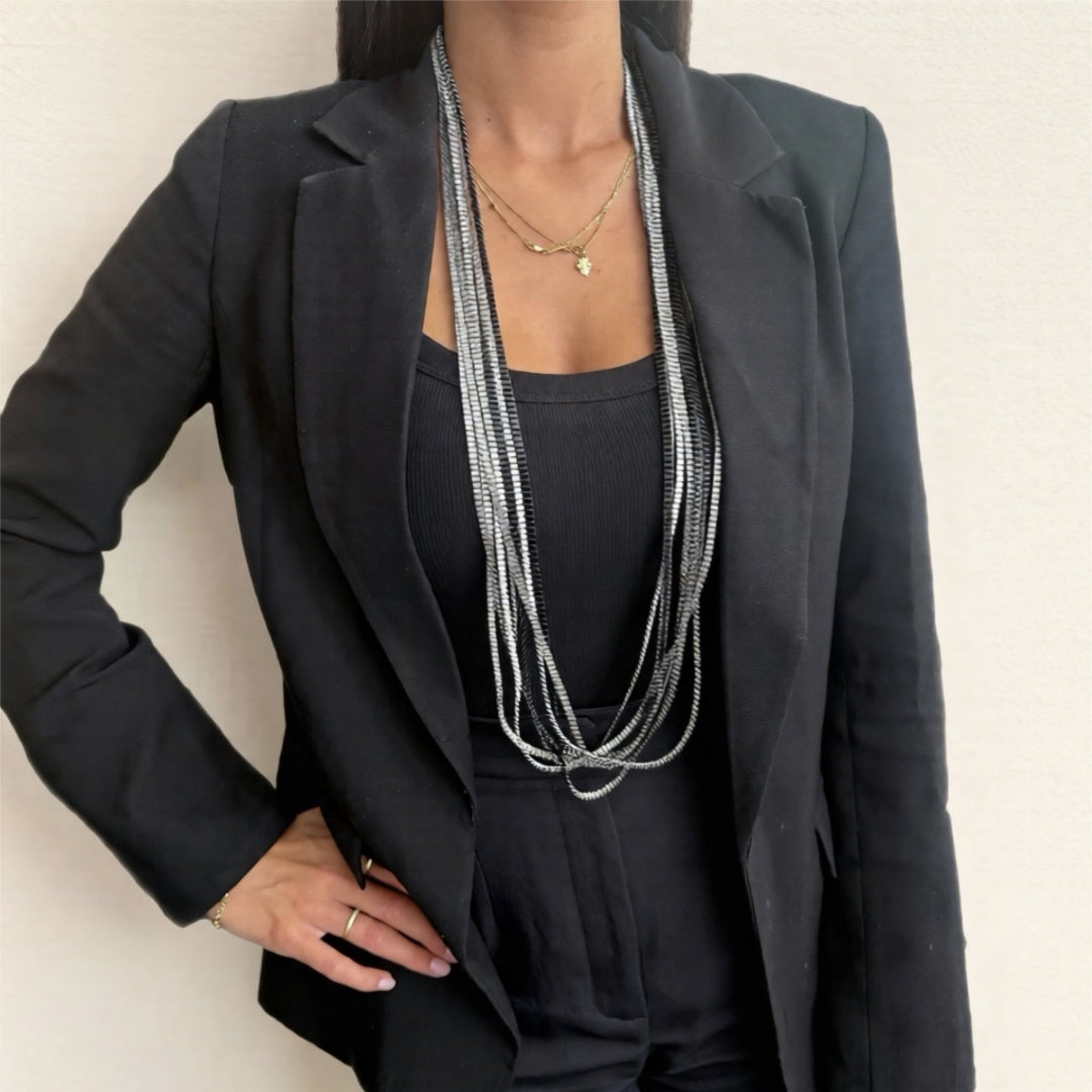 Person wearing a black suit with layered necklaces against a plain background