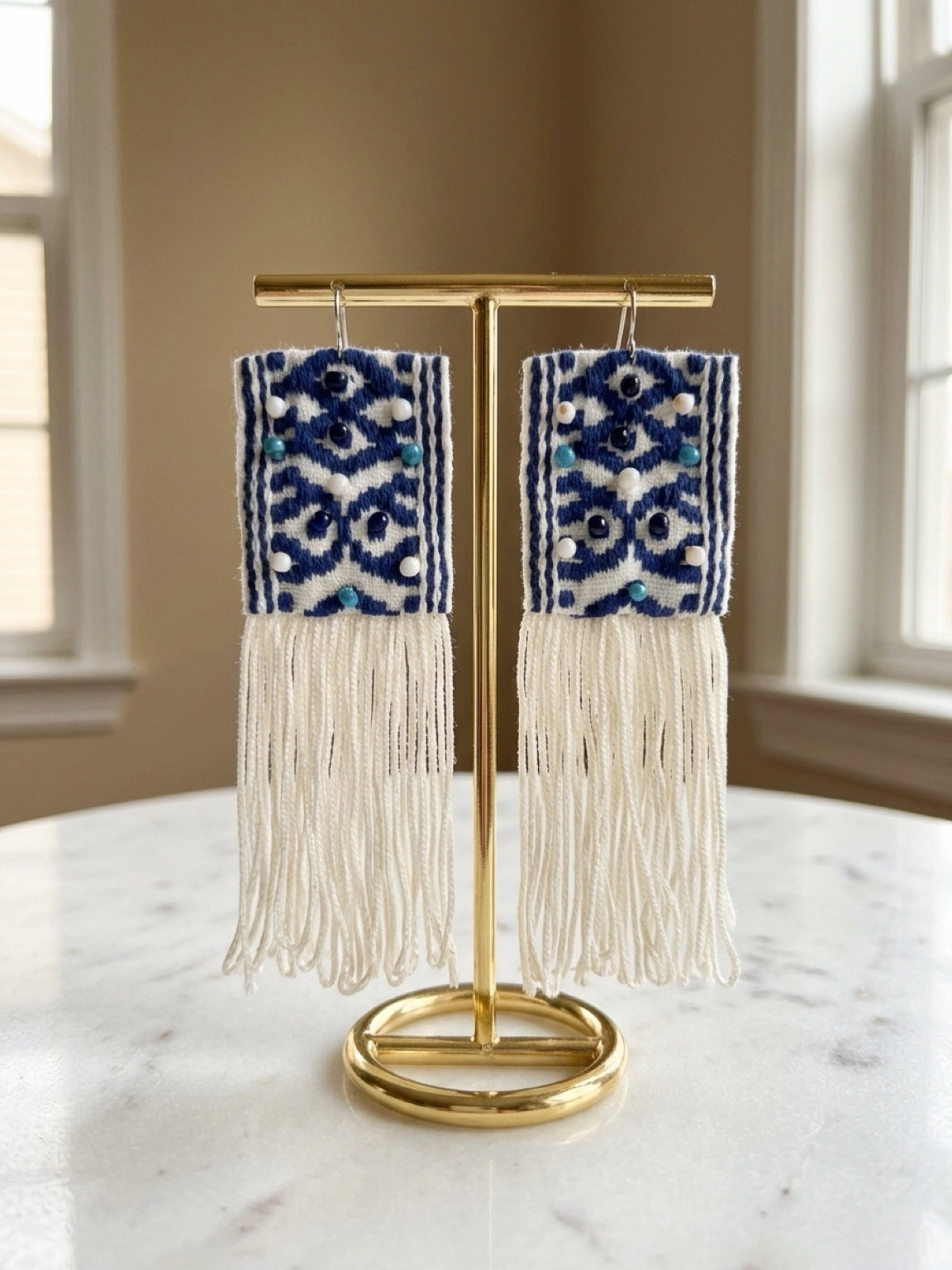 Boho-style rectangular dangle earrings with blue and white geometric patterns and long cream fringe, displayed on a gold stand on a marble table in a bright room.