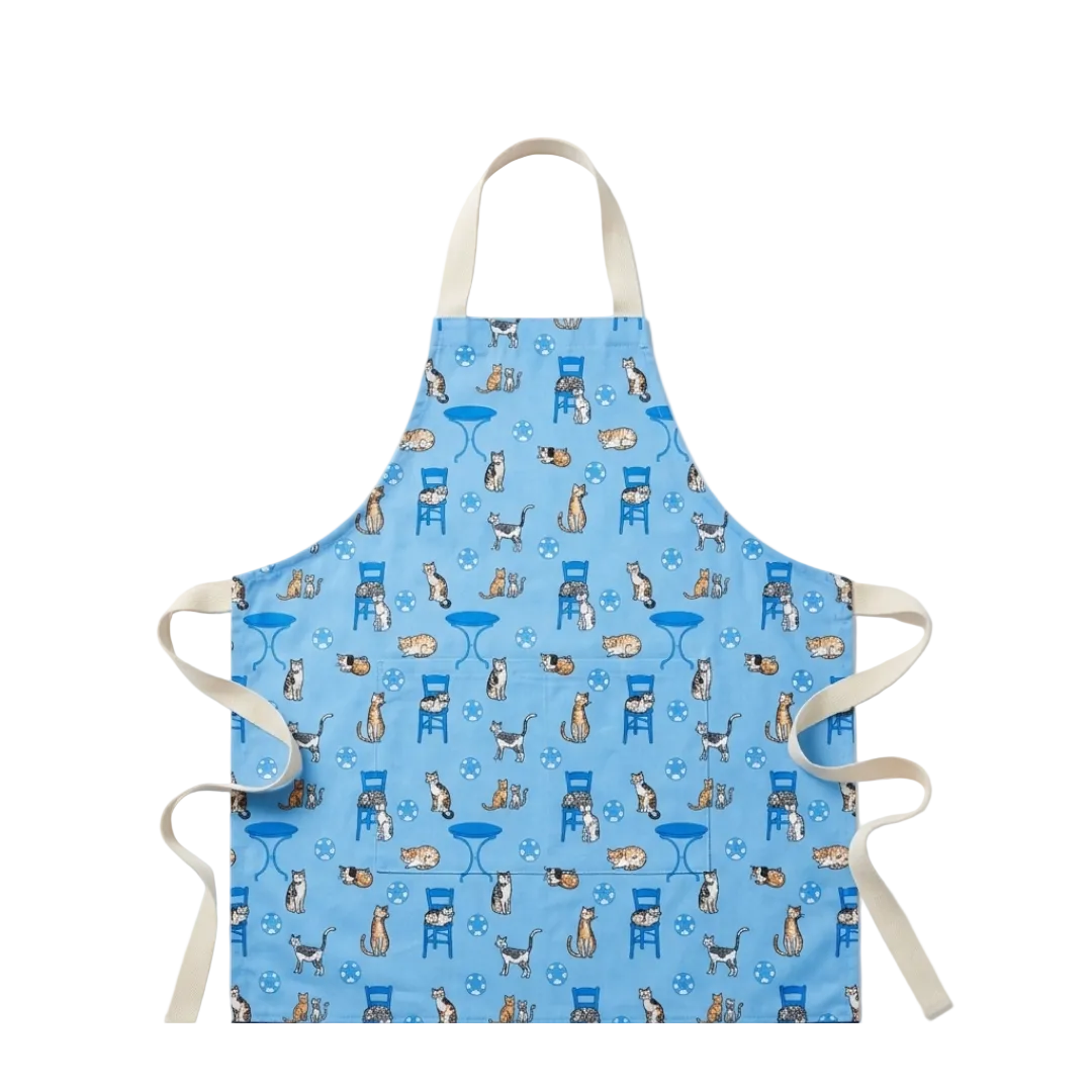 A light blue apron with a repeating pattern of cats sitting on chairs and tables, featuring cream-colored neck and waist straps, shown against a plain white background.