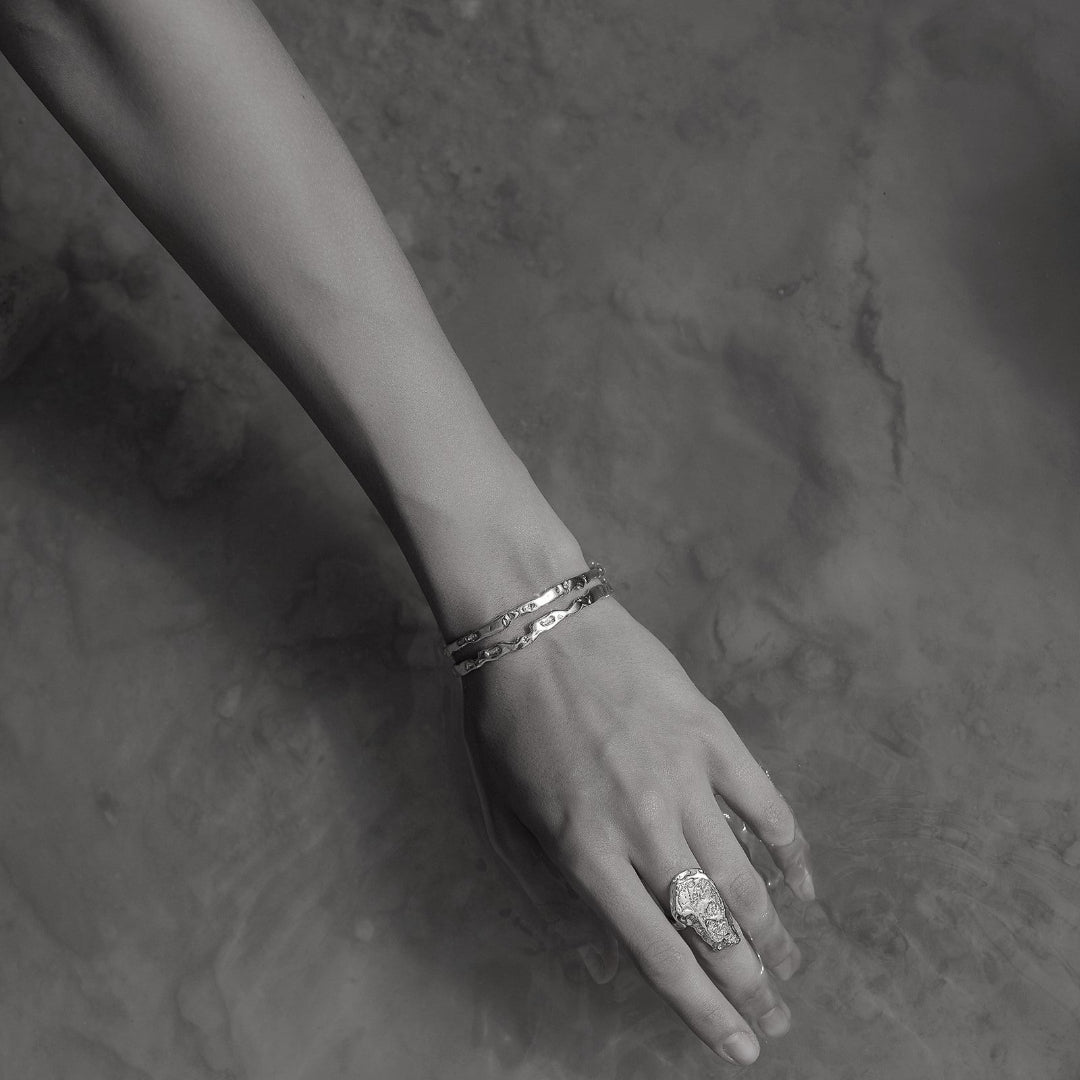 Black and white close-up of a hand with bracelets and a ring on a textured surface
