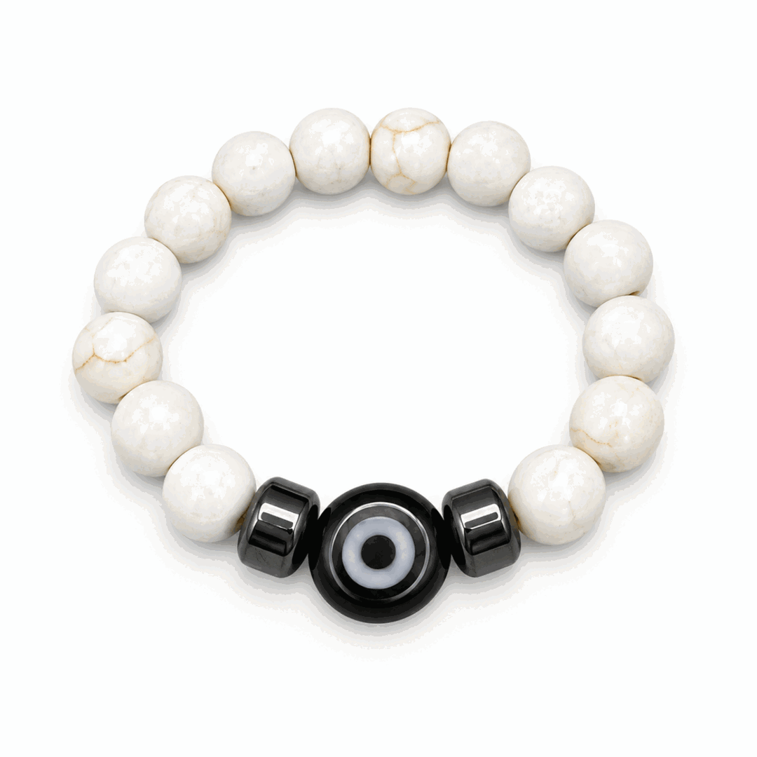 A beaded stretch bracelet featuring round off-white stone beads with natural veining, accented by two glossy black cylindrical beads and a central black evil eye charm with a white center, shown on a clean white background.