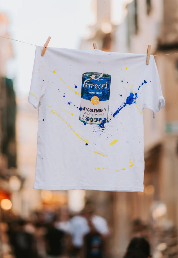White t-shirt with blue and yellow paint splashes and a soup can design, hanging on a line.