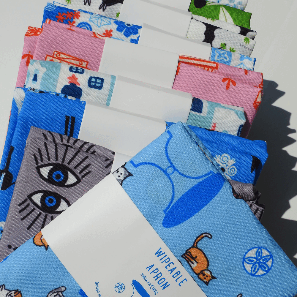 A bundle of folded fabric swatches with various colorful patterns, including blue with cartoon animals, pink with retro vehicles, gray with stylized eyes, and white with blue accents. A white paper label reading 'WIPEABLE APhON' is attached to the front blue fabric.