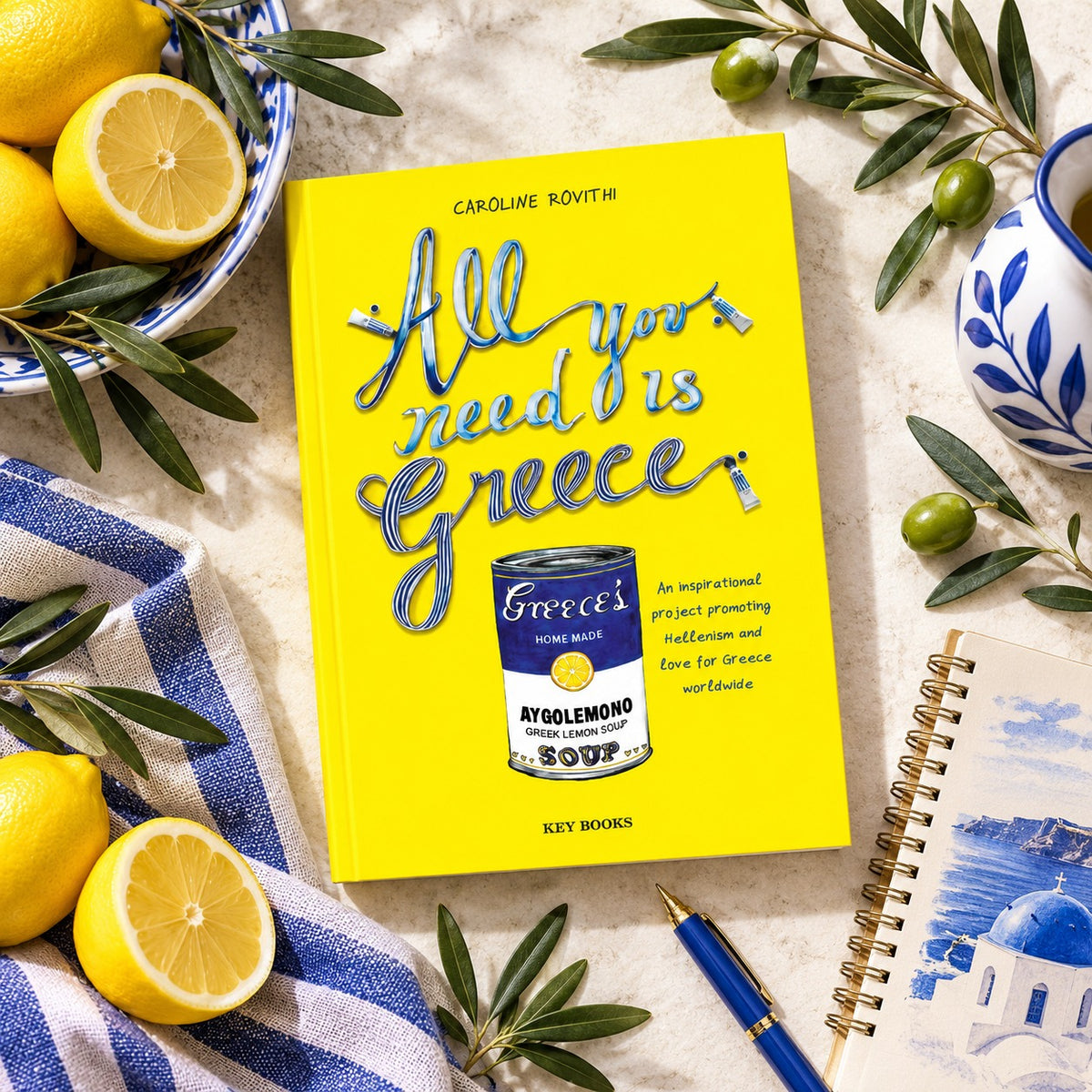 A bright yellow hardcover book titled "All you need is Greece" by Caroline Rovithi, styled in a flat lay with lemons, olives, a blue-and-white cloth, a blue-and-white ceramic vase, and a blue pen on a light stone surface.
