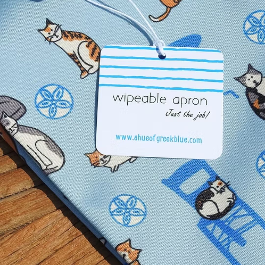 Greek Island Cats Apron – water-resistant apron with Greek cats, blue chairs and café tables on soft blue, detail view with wipeable apron tag | Thalassea