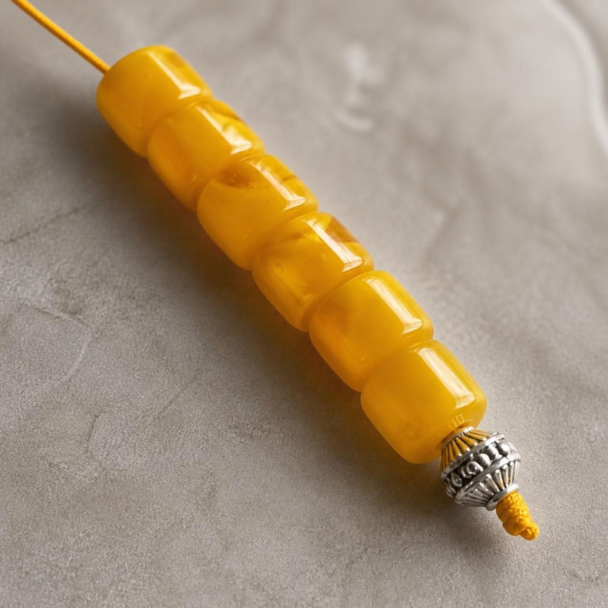 A close-up of a yellow beaded cord or necklace with a textured silver-toned end cap, photographed on a neutral gray surface.
