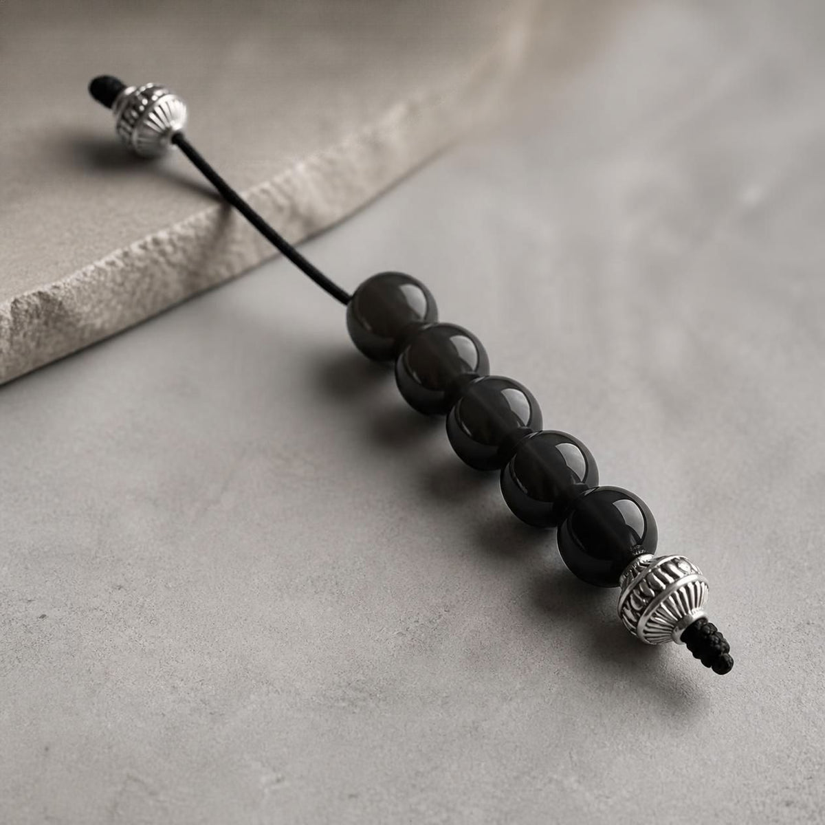 A beaded cord accessory with five glossy black round beads and textured silver metal end caps, photographed on a light gray surface with a stone slab prop.