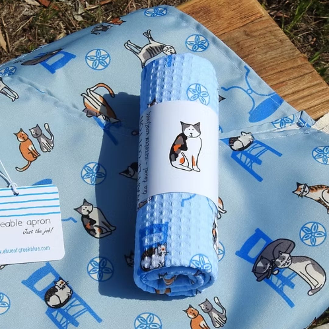 Greek Island Cats Apron – water-resistant apron with Greek cats, blue chairs and café tables on soft blue, styled with rolled tea towel | Thalassea