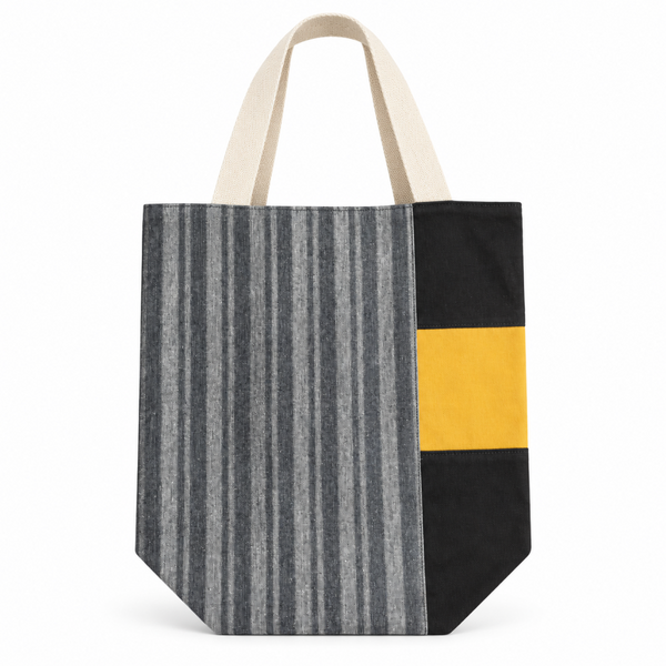 A canvas tote bag with cream-colored fabric handles, featuring a front panel split into a gray and black vertically striped section on the left and a black section with a wide mustard yellow band on the right.