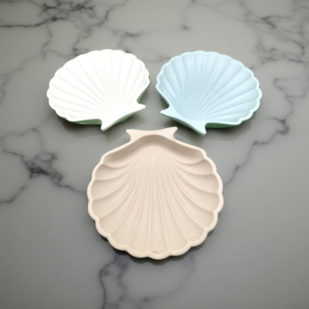 Three shell-shaped ceramic dishes in white, light blue, and beige on a marble surface.