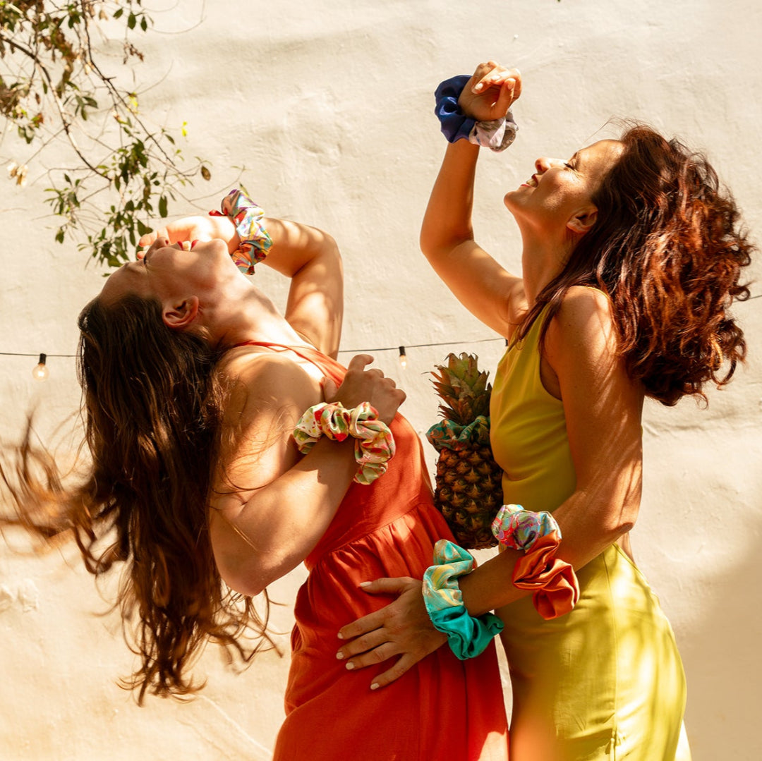 Two women in colorful outfits with a pineapple, dancing against a light background.