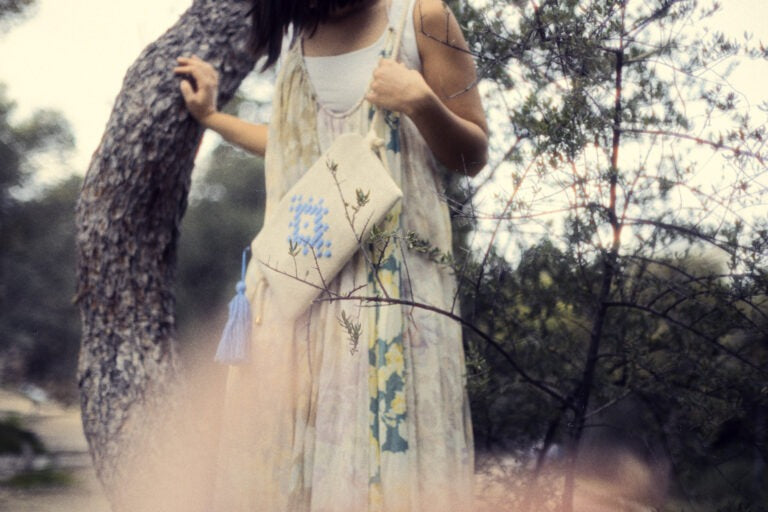 A woman in a bohemian-style cream dress stands outdoors next to a tree trunk, holding a matching cream tote bag with blue and green floral embroidery and a tassel.