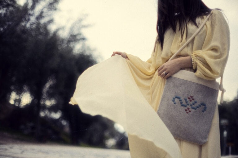 A woman in a pale yellow dress carries a large cream fabric bag outdoors. The bag has a gray front pocket with blue and red embroidered details and a white strap.