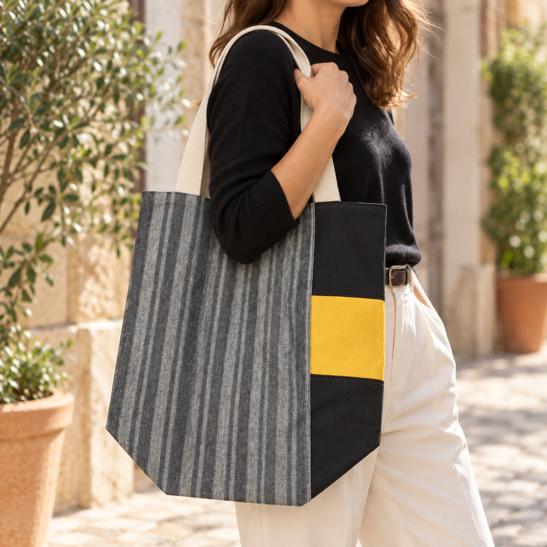 A woman carrying a large tote bag with a gray and white striped pattern, black side panels, and a bright yellow accent block, set against an outdoor stone walkway with potted plants.