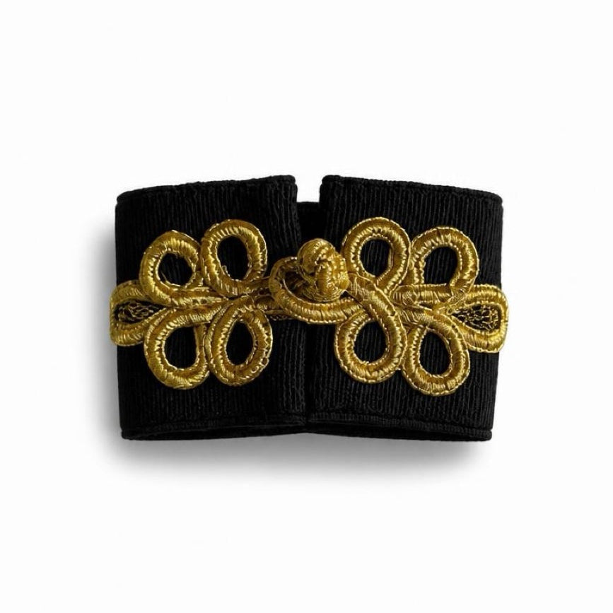 A black fabric cuff or wristband featuring a prominent gold embroidered decorative knot pattern across the front, displayed on a clean white background.