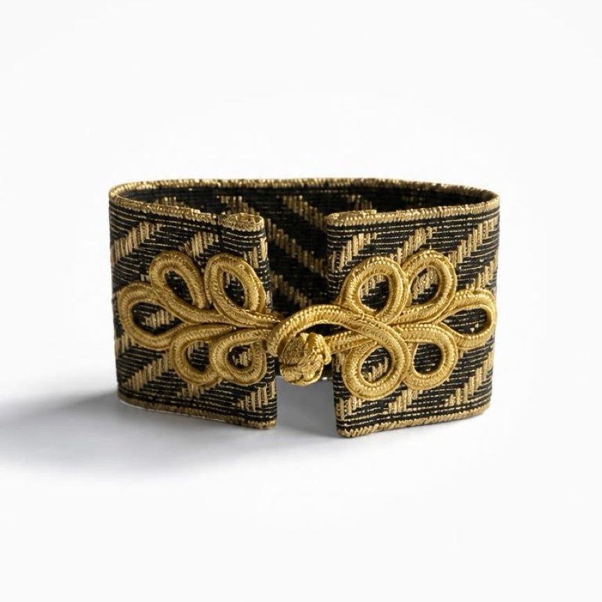 A wide cuff bracelet featuring a black and gold woven pattern with a large gold decorative knot design on the front, set against a plain white background.