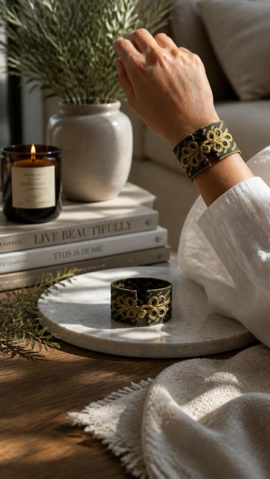 A person wearing a wide black cuff bracelet with an intricate gold floral pattern, shown in a cozy, styled home environment with a matching bracelet resting on a marble tray.