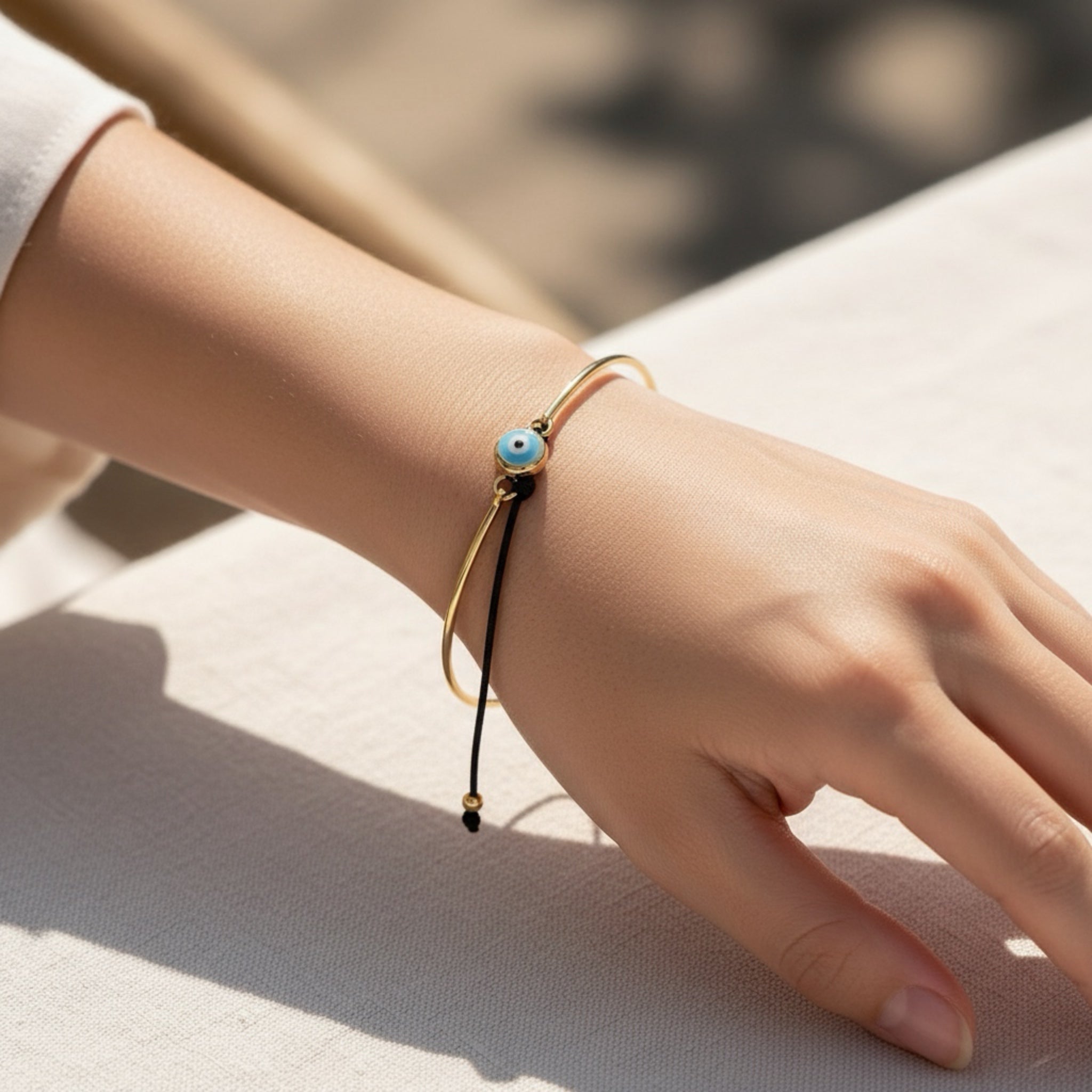 Close-up of a hand wearing a gold bracelet with a blue bead on a light surface.
