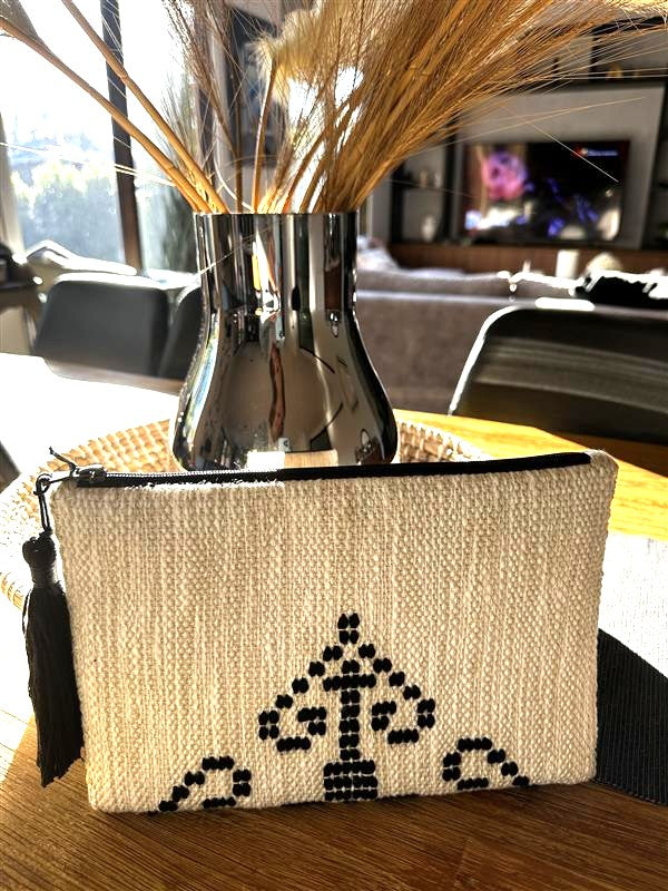 Woven pouch with decorative design on a table, next to a metallic vase with dried plants.