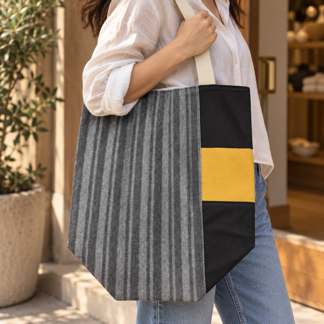 A person wearing a white button-up shirt and blue jeans carries a large tote bag with a gray vertically striped panel, a black panel, and a mustard yellow accent panel on the side.