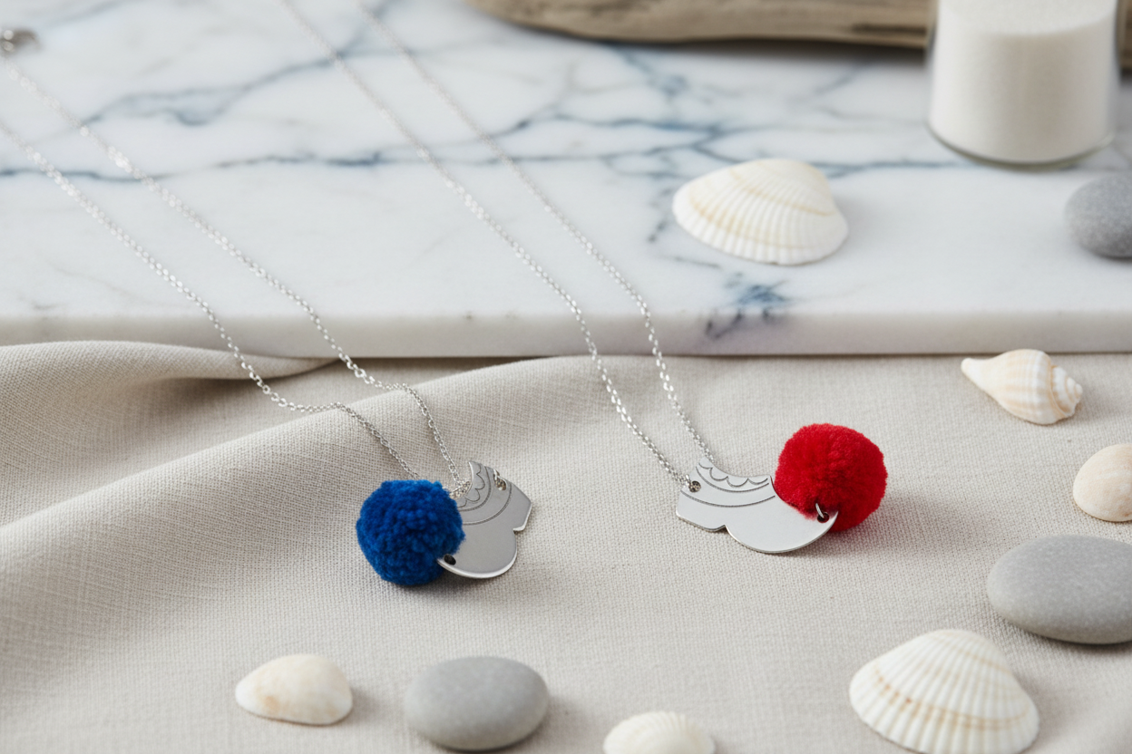 Two necklaces with colorful pom-poms on a marble surface with seashells.