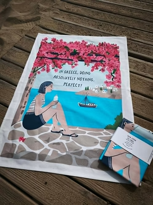 In Greece, Doing Absolutely Nothing. Perfect! Cotton Tea Towel – woman under pink bougainvillea gazing at a turquoise bay, fishing boat and white island village, styled with card-wrapped towel | Thalassea